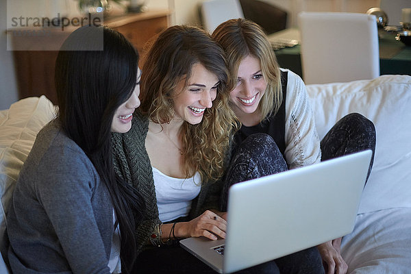 Drei fröhliche Frauen sitzen auf der Couch und schauen zusammen auf einen Laptop