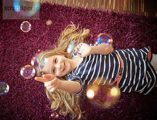 Glückliches Mädchen liegt auf einem Teppich und spielt mit Seifenblasen