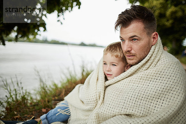 Vater und Sohn sitzen am Flussufer eingewickelt in eine Decke