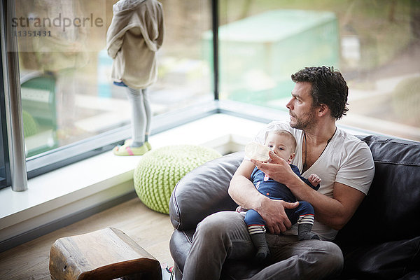 Vater gibt seinem Baby das Fläschchen auf der Couch mit Tochter am Fenster