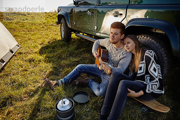 Junges Paar sitzt auf einer Wiese neben einem Auto und macht Musik