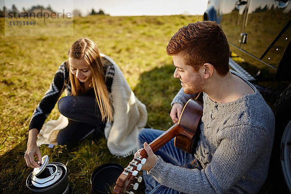 Junges Paar sitzt auf einer Wiese neben einem Auto und einem Campingkocher und macht Musik