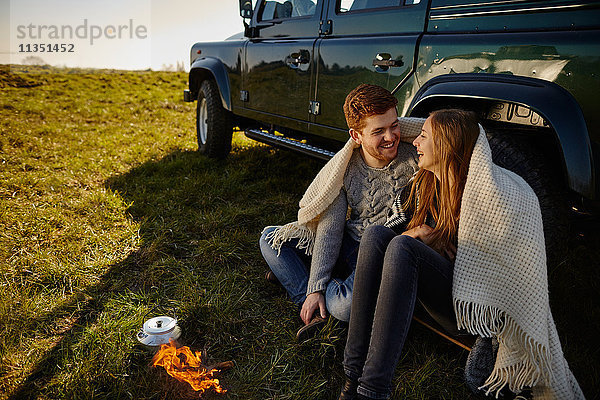 Glückliches junges Paar sitzt auf einer Wiese neben einem Auto und Campingkocher