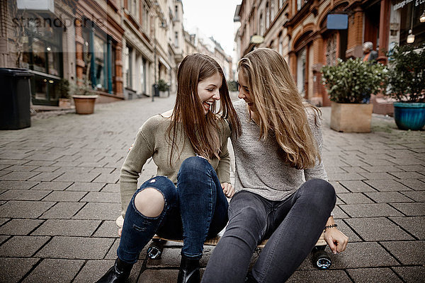 Zwei fröhliche junge Frauen sitzen auf einem Skateboard in der Stadt