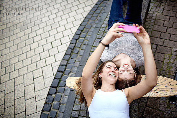 Zwei fröhliche junge Frauen liegen auf einem Skateboard und machen ein Selfie