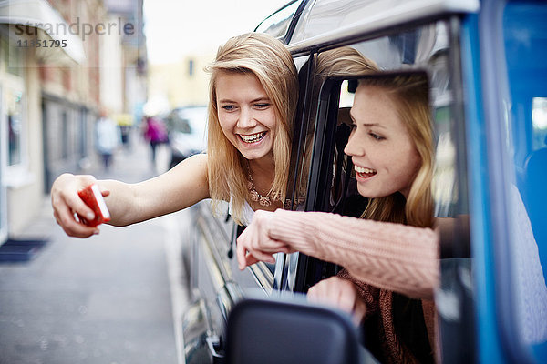 Zwei fröhliche junge Frauen lehnen sich aus dem Autofenster und machen ein Selfie