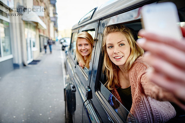 Zwei fröhliche junge Frauen lehnen sich aus dem Autofenster und machen ein Selfie