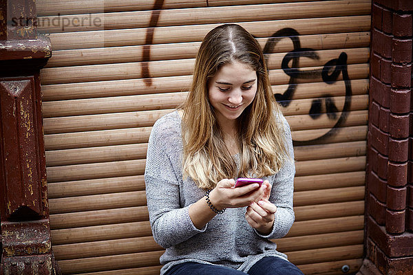 Lächelnde junge Frau  die auf der Türschwelle sitzt und ihr Mobiltelefon überprüft