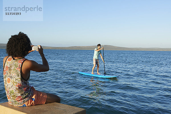 Junger Mann fotografiert Freund beim Paddeln auf dem sonnigen Sommermeer