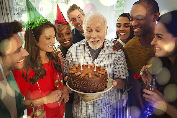 Multi-ethnische Familie beobachtet Senior Mann blasen Geburtstag Kerzen auf Schokolade Kuchen