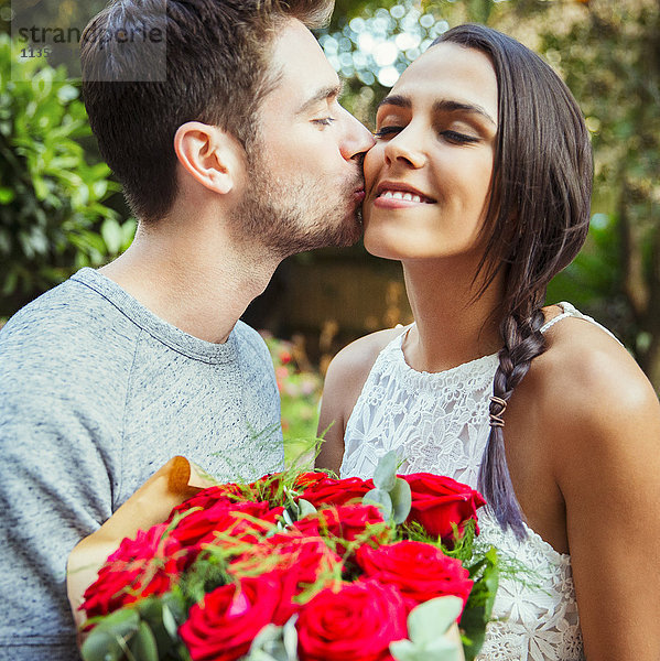 Zärtlicher Mann  der der Freundin einen roten Rosenstrauß schenkt und sie auf die Wange küsst.