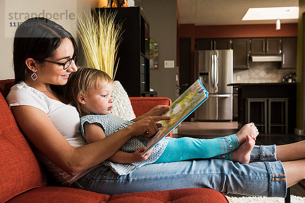 Mutter lehrt Tochter  das Buch auf dem Sofa zu lesen.