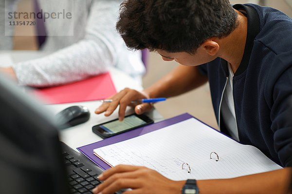 Junge männliche College-Studenten am Computerschreibtisch beim Rechnen mit einem Smartphone