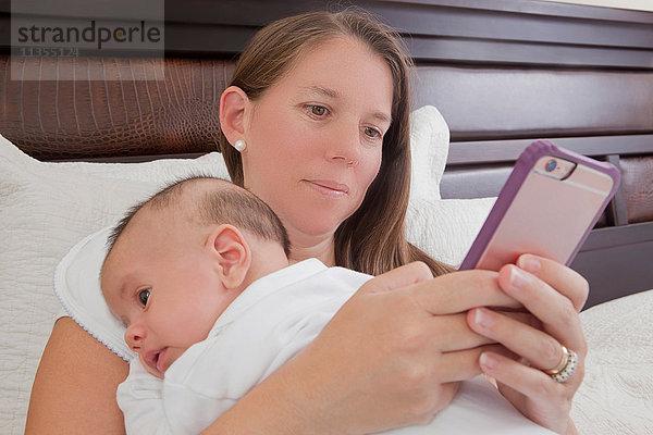Mutter im Bett hält Baby Junge  der mit Smartphone simst