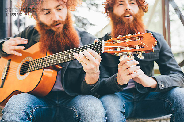 Junge männliche Hipster-Zwillinge mit roten Bärten beim Gitarrespielen sitzend