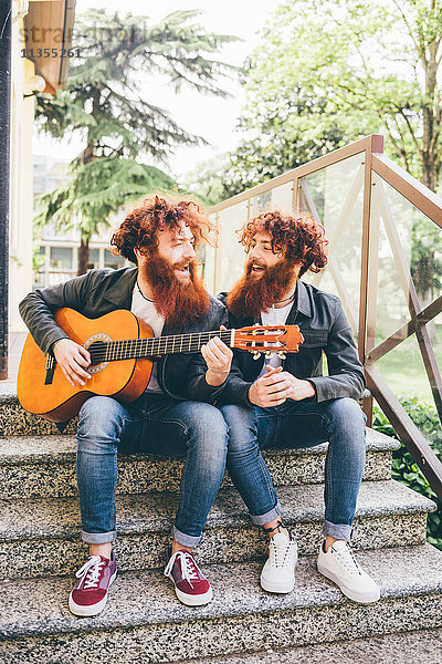 Junge männliche Hipster-Zwillinge mit roten Bärten sitzen auf einer Treppe und spielen Gitarre