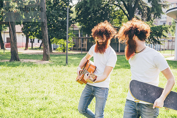 Junge männliche Hipster-Zwillinge mit roten Haaren und Bärten spazieren im Park und spielen Gitarre