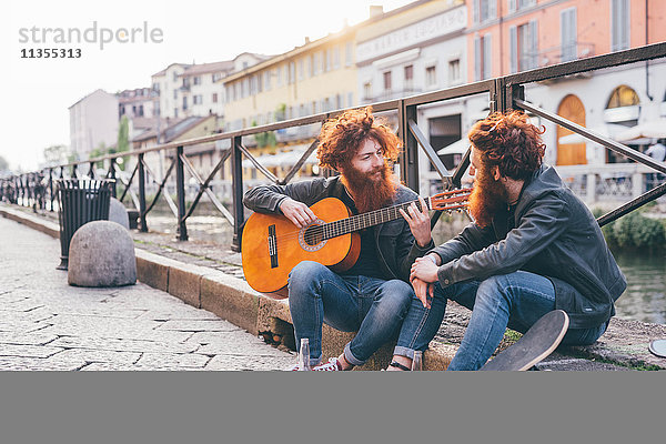 Junge männliche Hipster-Zwillinge mit roten Haaren und Bärten spielen Gitarre am Kanalufer.