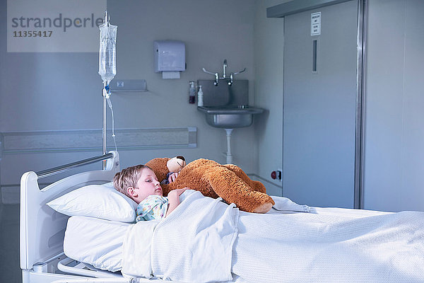 Junge Patientin im Bett umarmt Teddybär auf Krankenhaus-Kinderstation