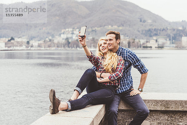 Junges Paar sitzt auf der Hafenmauer und nimmt Smartphone-Selfie mit  Comer See  Italien