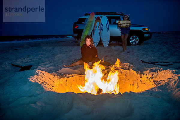 Junges Mädchen sitzt am Lagerfeuer am Strand  ihre Schwester trägt Feuerholz zum Feuer