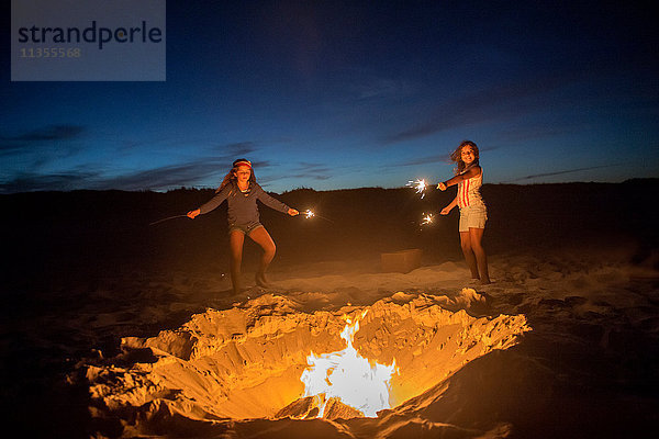 Zwei junge Mädchen halten am Strand am Lagerfeuer Wunderkerzen und halten Wunderkerzen