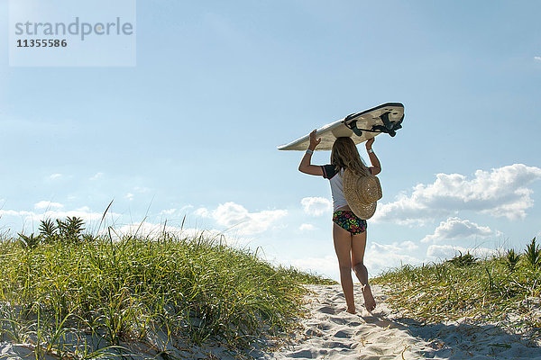 Junges Mädchen geht auf Sanddünen  trägt ein Surfbrett  Rückansicht