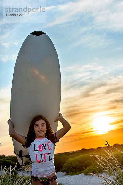 Junges Mädchen am Strand  ein Surfbrett tragend  bei Sonnenuntergang