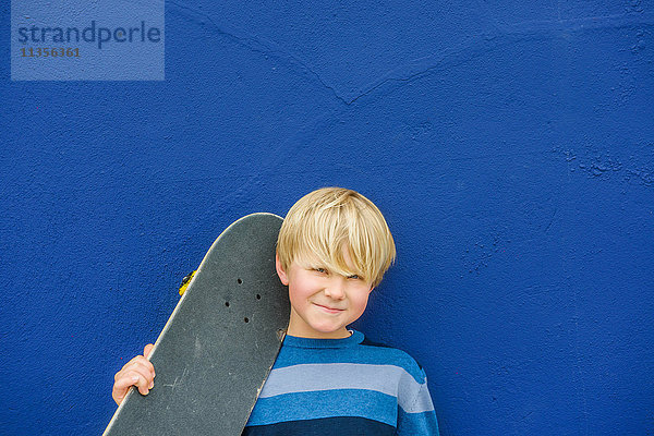 Porträt eines süßen Jungen  der ein Skateboard auf der Schulter trägt  vor einer blauen Wand