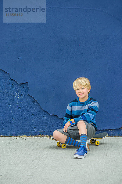 Porträt eines Jungen  der auf einem Skateboard vor einer blauen Wand sitzt