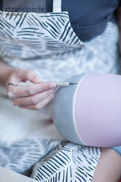Nahaufnahme der Hände einer Töpferin  die in der Werkstatt Glasur auf eine Vase malt
