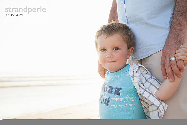 Junge am Strand  der die Hände seines Vaters hält und lächelnd in die Kamera schaut