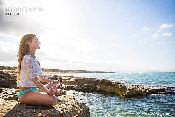 Junge Frau sitzt auf Felsen am Meer  in Yogastellung