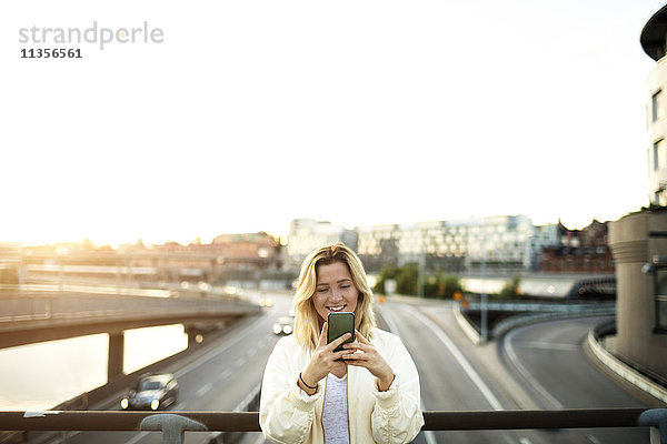 Fröhliche junge Frau mit dem Handy auf der Brücke gegen den Himmel stehend