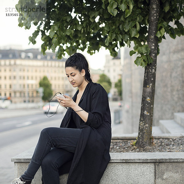 Junge Frau  die ein Smartphone benutzt  während sie auf einer Stützmauer in der Stadt sitzt.