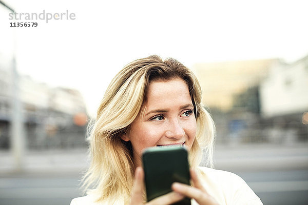 Glückliche junge Frau mit Handy in der Stadt gegen den klaren Himmel