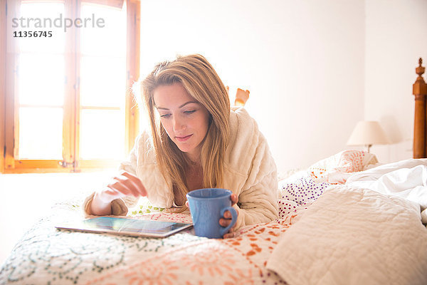 Junge Frau liegt im Bett und stöbert morgens auf einem digitalen Tablet