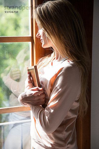 Junge Frau umarmt Bilderrahmen und blickt aus dem Fenster