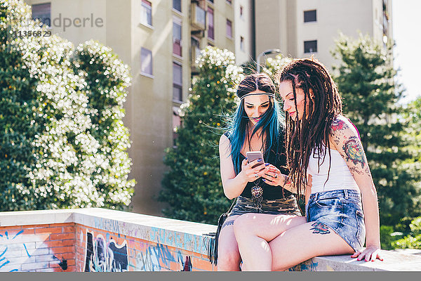 Zwei junge Frauen an der Wand beim Lesen von Smartphone-Texten in einer städtischen Wohnsiedlung