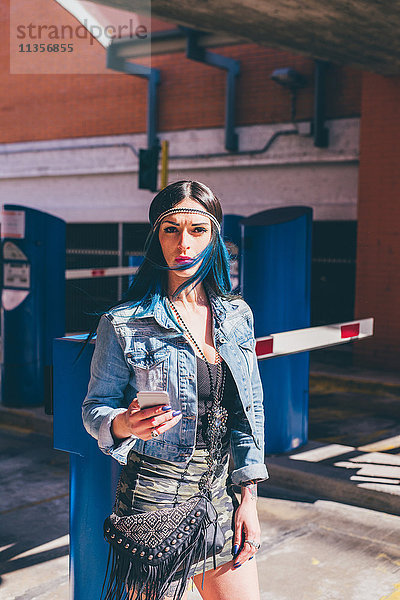 Junge Frau mit tauchgefärbten blauen Haaren mit Smartphone in einer städtischen Wohnsiedlung