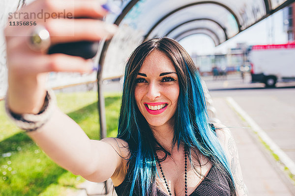 Junge Frau mit tauchgefärbten blauen Haaren nimmt Smartphone im städtischen Buswartehäuschen