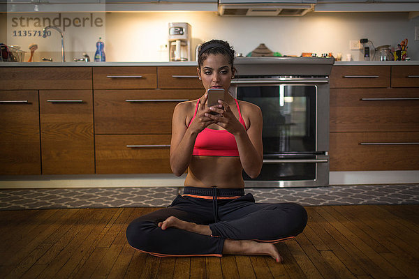 Junge Frau in der Küche  sitzt in Yoga-Position und benutzt ein Smartphone