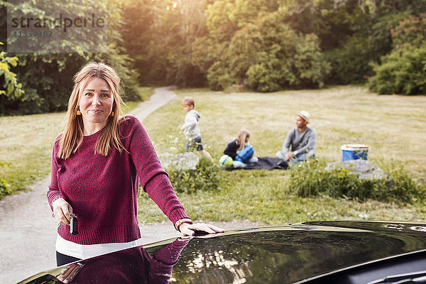 Porträt einer fröhlichen Frau am Elektroauto mit Familie im Hintergrund im Park