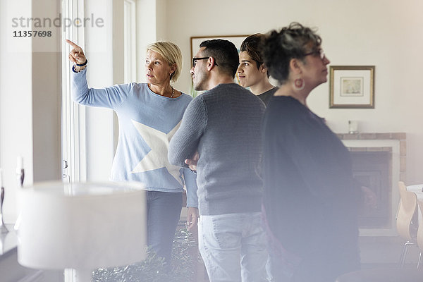 Reife Frau  die durchs Fenster zeigt und Freunden zeigt  während sie im Wohnzimmer steht.