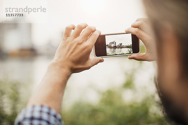 Beschnittenes Bild eines Mannes  der ein Silo durch ein Smartphone gegen den Fluss fotografiert.