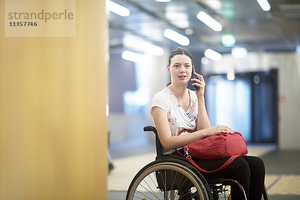 Porträt einer jungen Frau  die im Rollstuhl auf einem Smartphone spricht