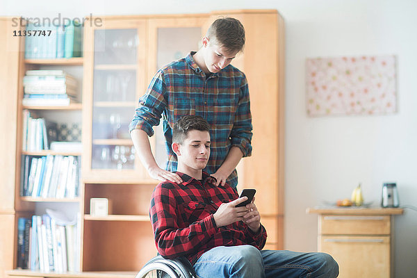 Junger Mann im Rollstuhl beim Lesen von Smartphone-Texten in der Küche