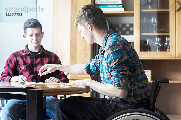Junger Mann im Rollstuhl beim Damespiel mit einem Freund in der Küche