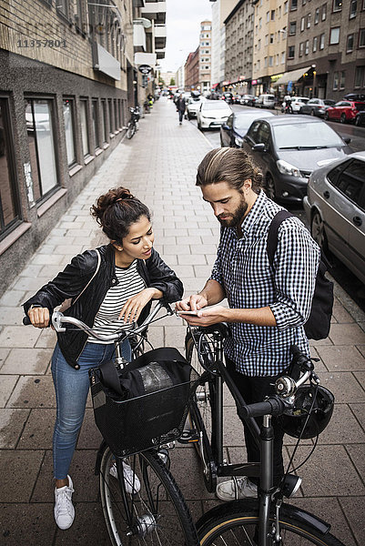 Hochwinkelansicht von Freunden mit Fahrrädern  die auf dem Bürgersteig in der Stadt stehen und das Telefon anschauen