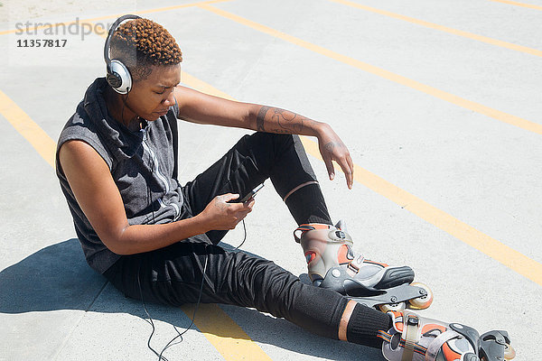 Frau mit Rollerblades und Kopfhörern sitzt auf dem Boden und schaut auf ein Smartphone